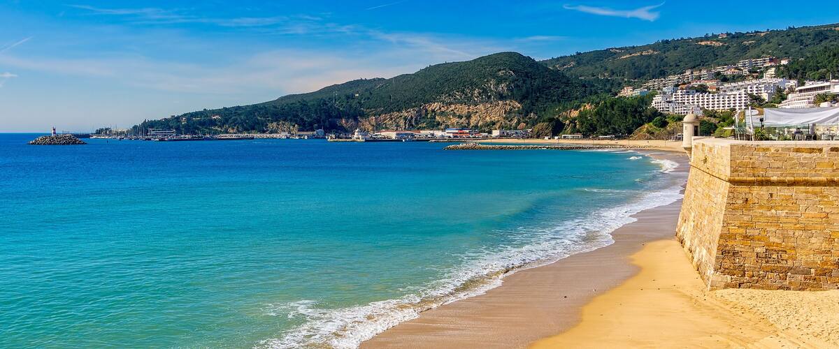 A coastal scene with a sandy beach, a stone wall, and a backdrop of hills and buildings.