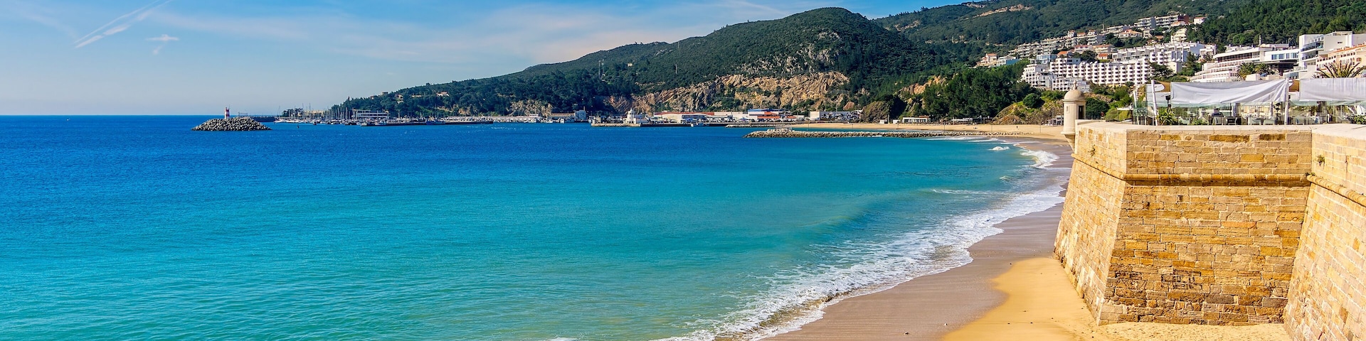 A coastal scene with a sandy beach, a stone wall, and a backdrop of hills and buildings.