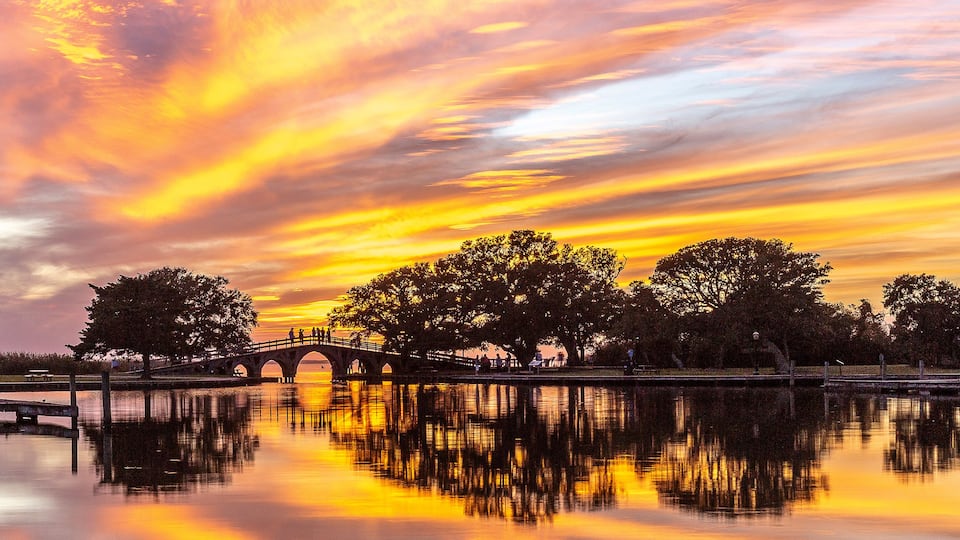 A sunset over a calm lake with a bridge and trees reflecting on the water.