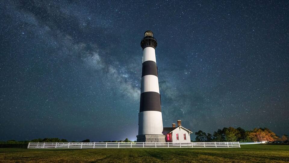 A lighthouse with alternating black and white stripes, a small adjacent building, and a white fence in a grassy field under a starry night sky.