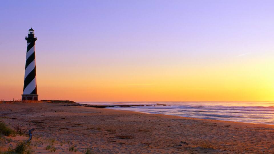 A lighthouse on a beach at sunset with the ocean in the foreground.