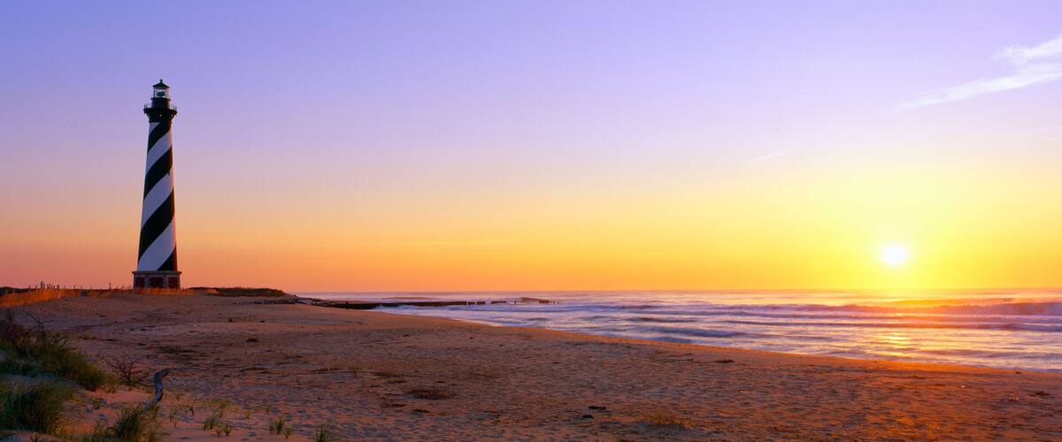 A lighthouse on a beach at sunset with the ocean in the foreground.