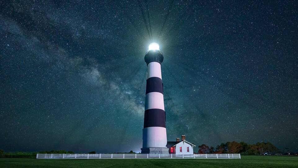 A lighthouse with alternating black and white stripes, a white fence, and a small red building at its base.