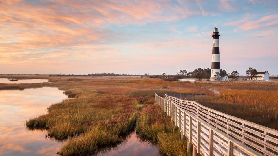 A lighthouse, a wooden boardwalk, and a marshy landscape with a sunset sky.