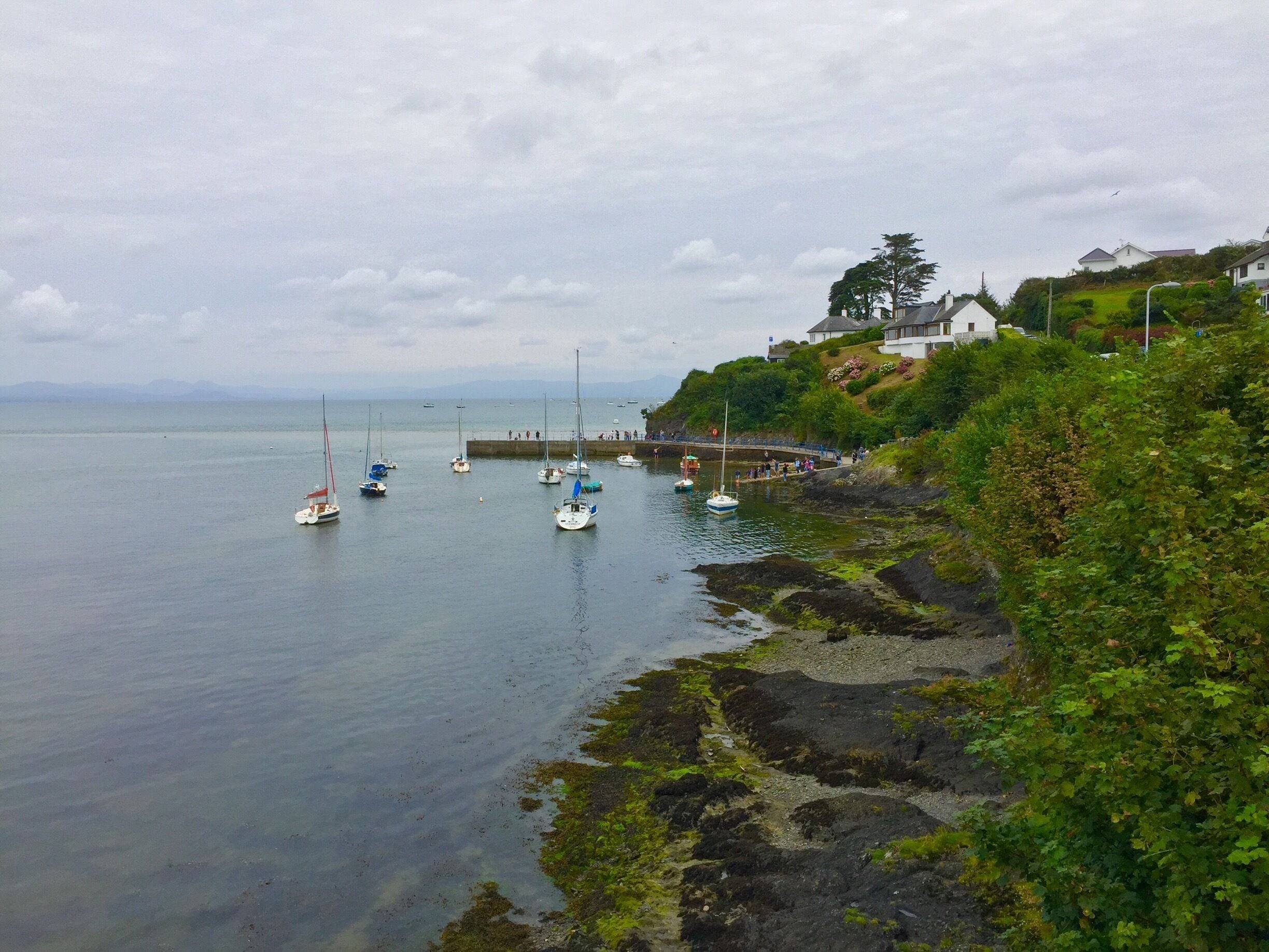 A coastal scene with sailboats anchored near a rocky shore, a promenade with people, and houses on a hillside.