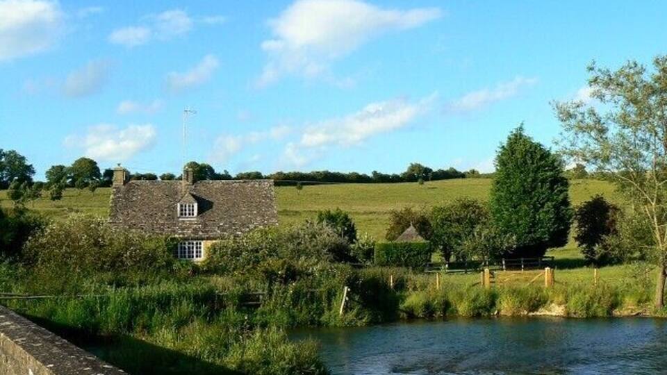 A stone house by a river with a wooden fence and a thatched-roof structure in the background.