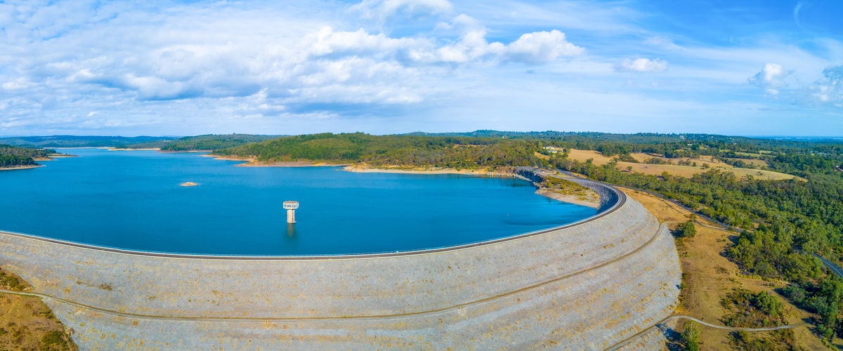 A large reservoir with a dam, surrounded by forested hills and a clear sky.