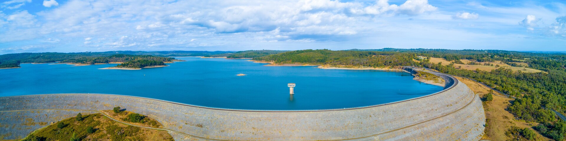 A large reservoir with a dam, surrounded by forested hills and a clear sky.