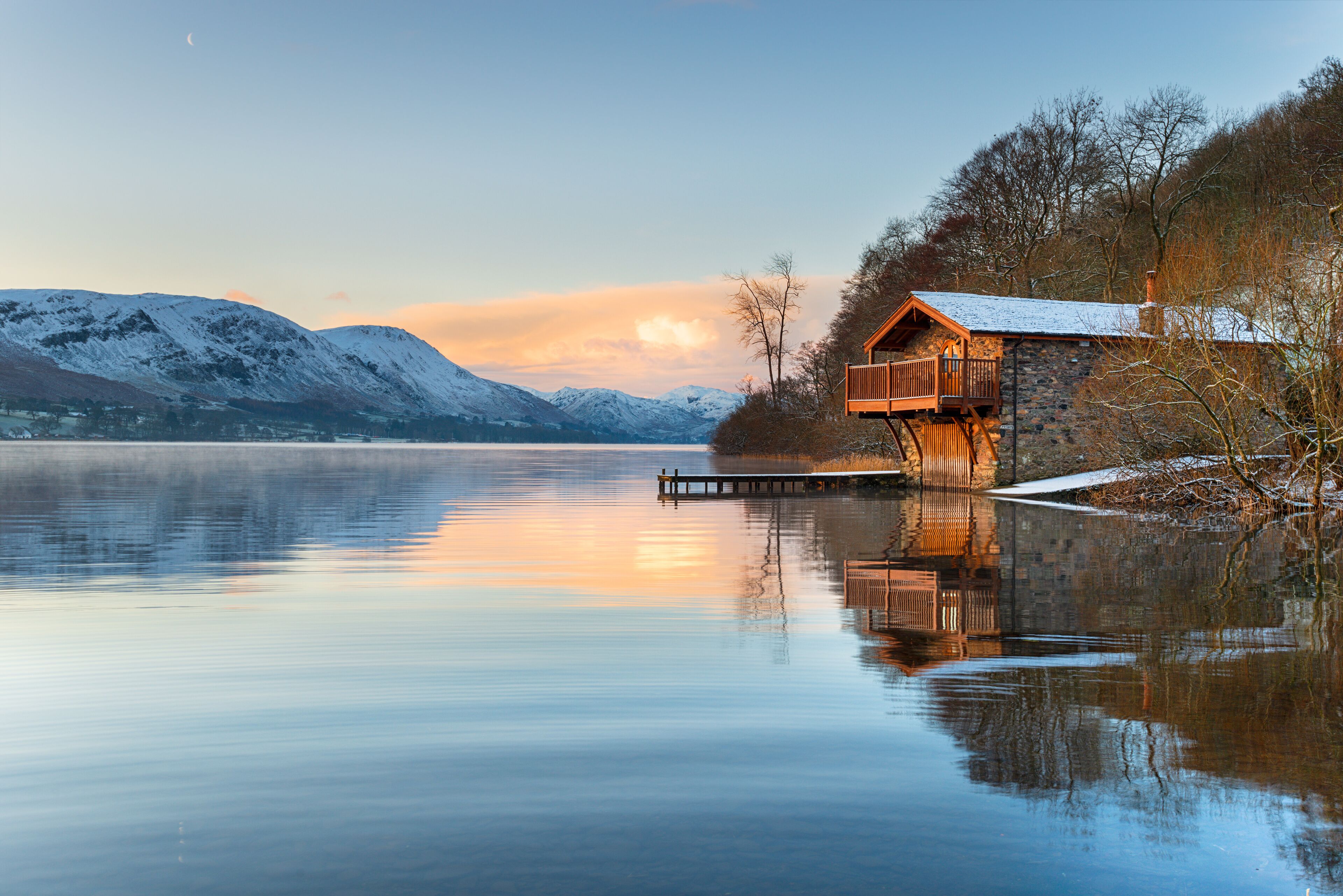 A wooden cabin with a balcony on a lakeside, surrounded by snow-covered mountains and a calm lake.