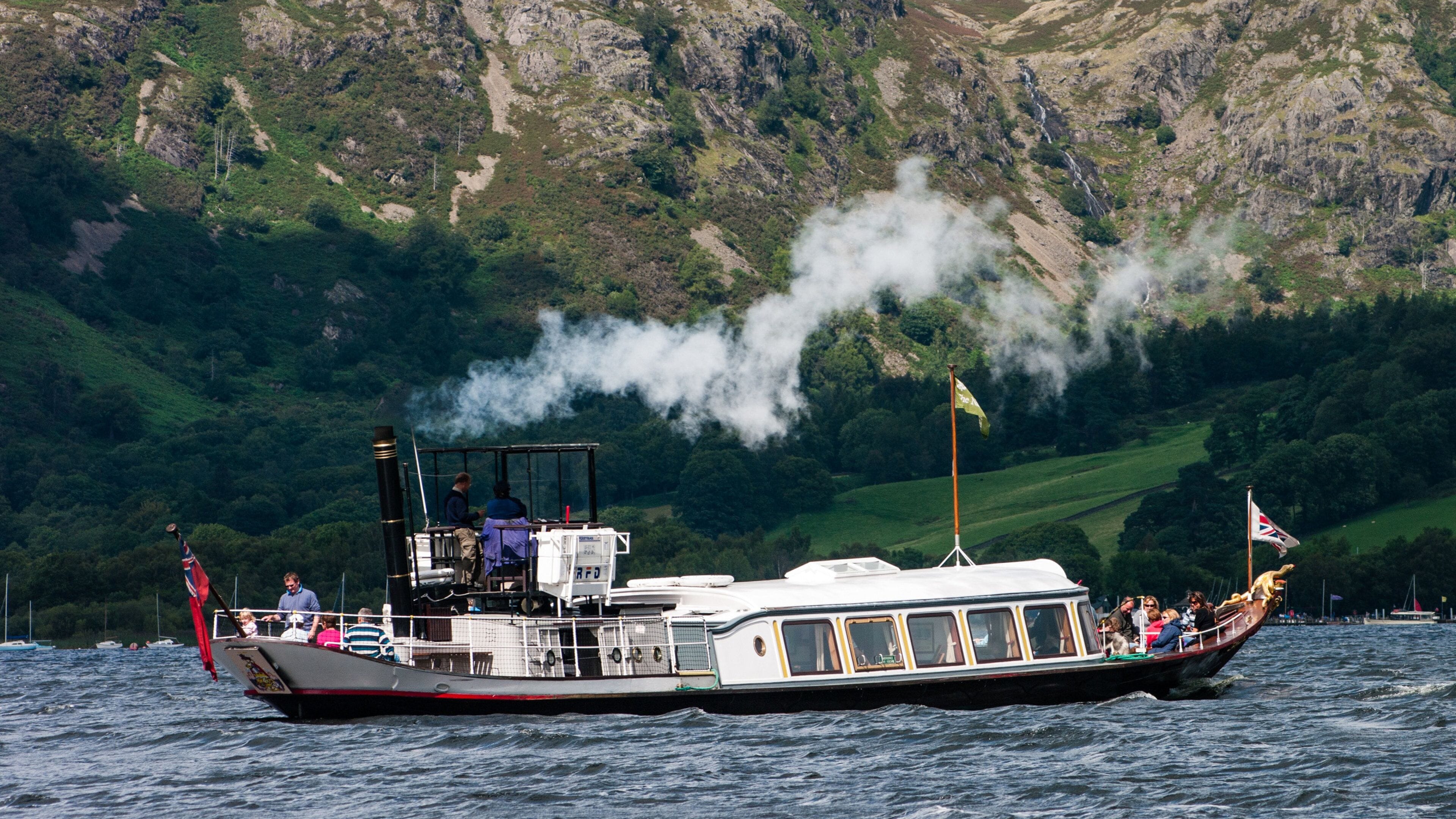 A steamboat with passengers and a flag, surrounded by a mountainous landscape and a body of water.