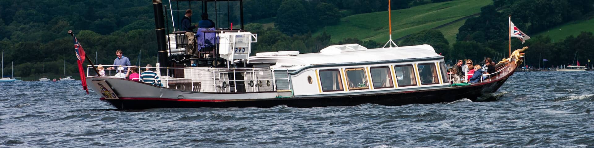 A steamboat with passengers and a flag, surrounded by a mountainous landscape and a body of water.