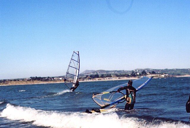 Two windsurfers on the water with a beach and hills in the background.