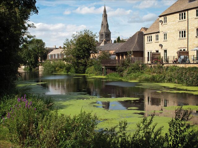 A river with green algae, a church steeple, and stone buildings along the riverbank.
