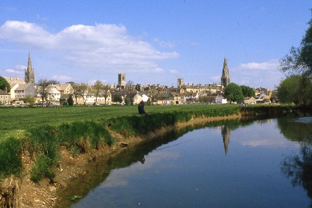 A river with a person walking along the bank, a town with several buildings and spires in the background, and a clear blue sky.