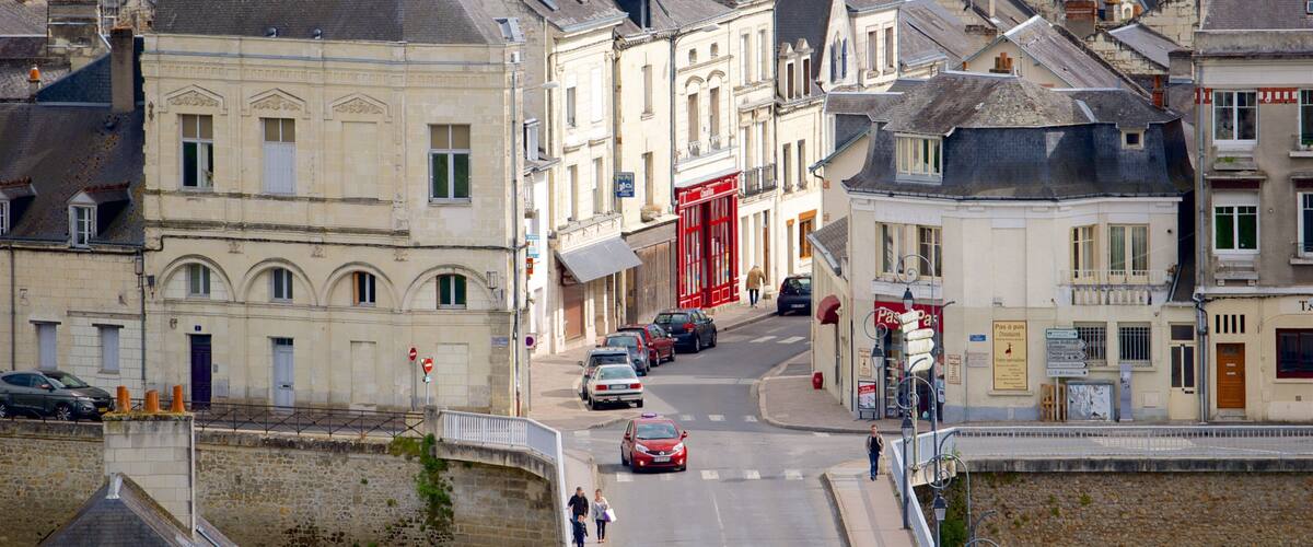 A street in a European town with historic buildings, cars parked along the road, and pedestrians walking.