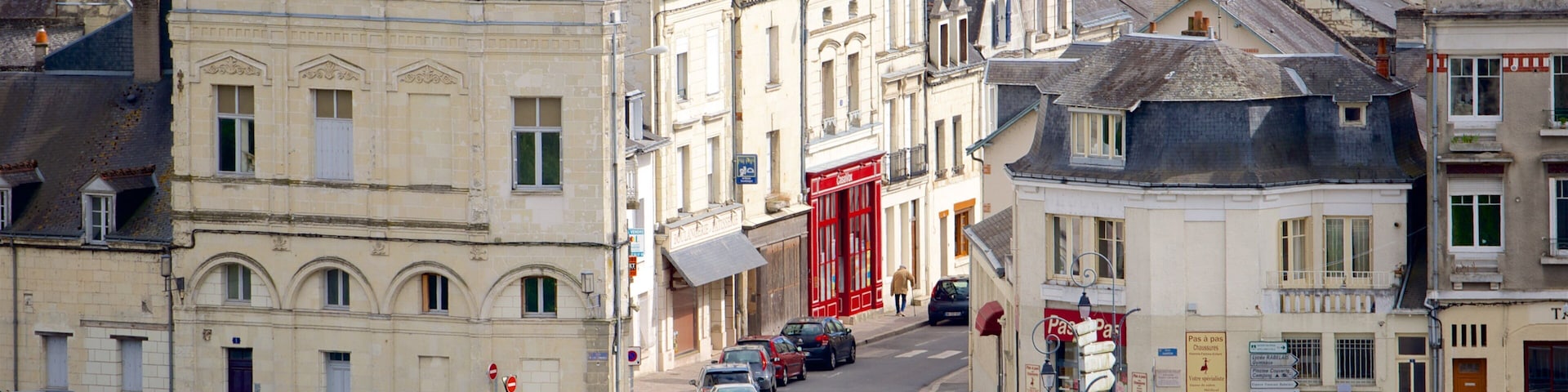 A street in a European town with historic buildings, cars parked along the road, and pedestrians walking.