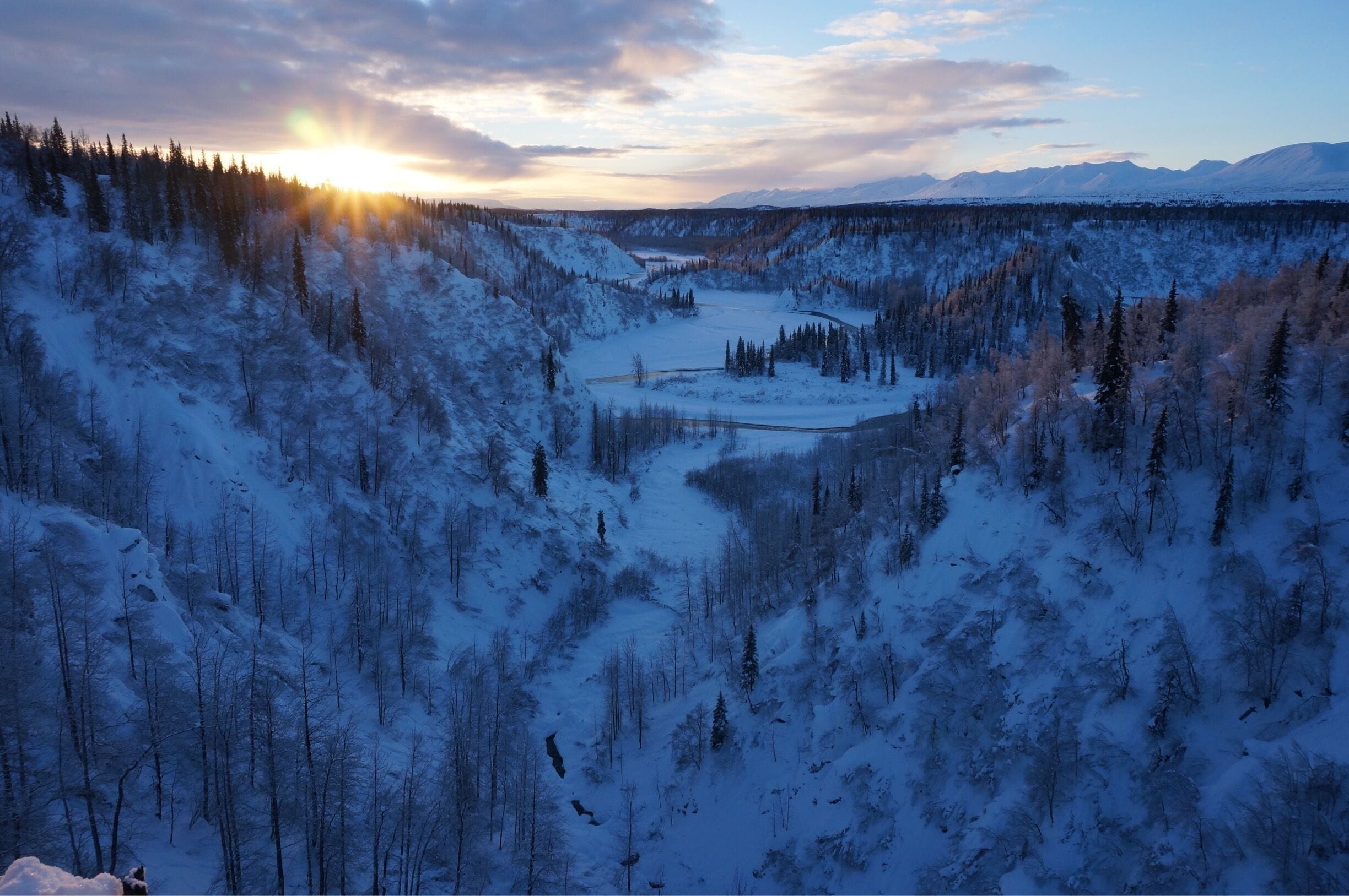 A snow-covered valley with a river, dense forest, and mountains in the background.
