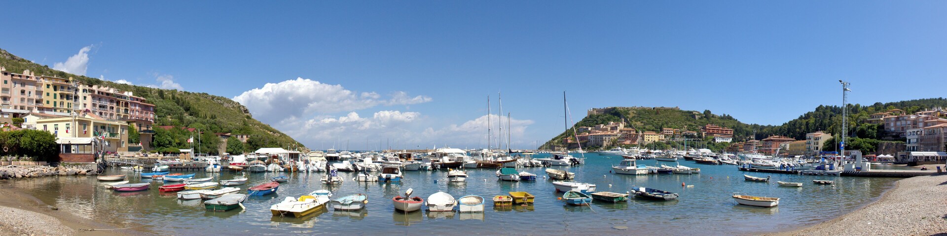 A marina filled with small boats, a rocky shoreline, and buildings on the hills in the background.