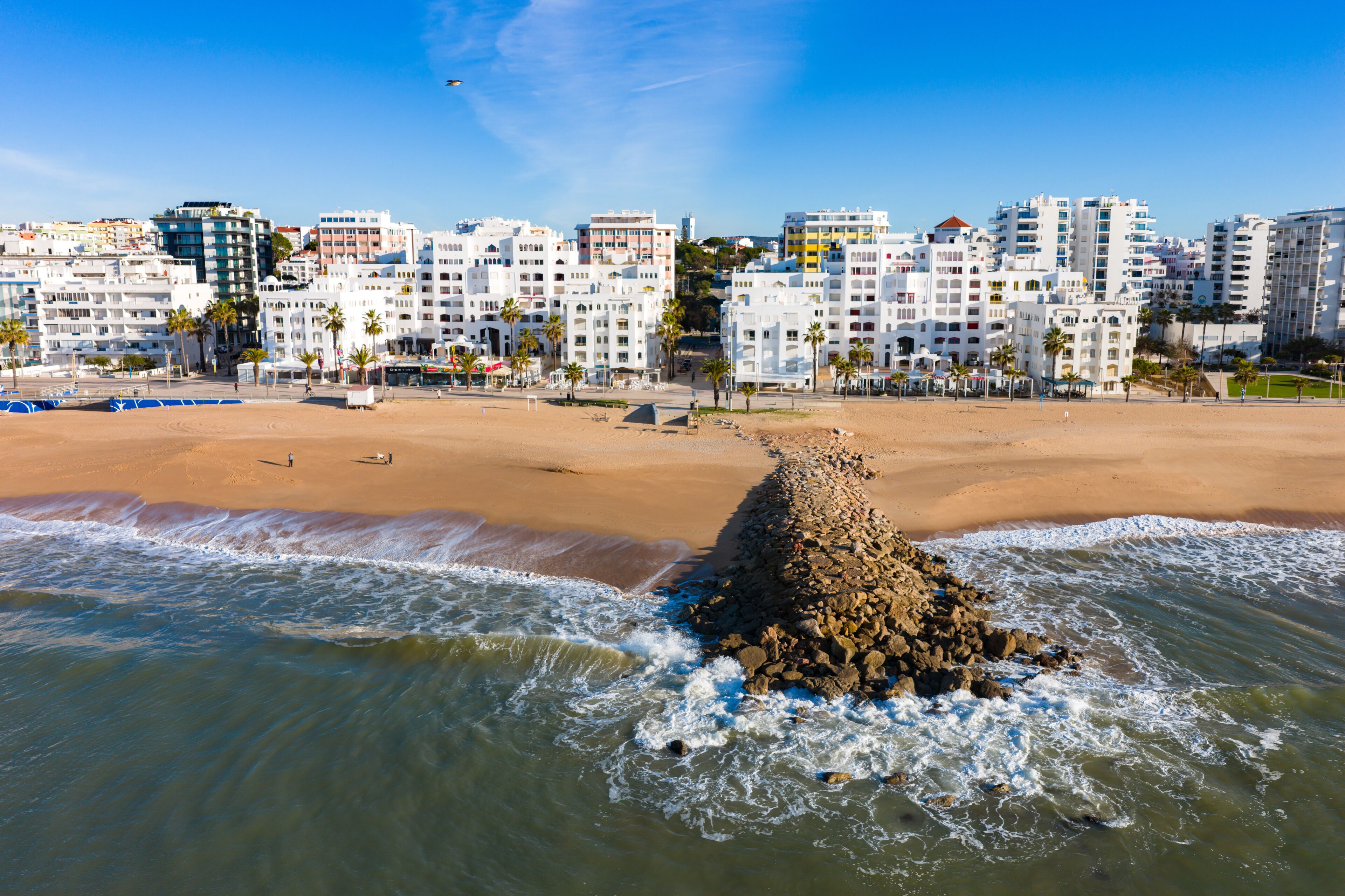 A coastal cityscape with a sandy beach, a rocky breakwater, and modern high-rise buildings.