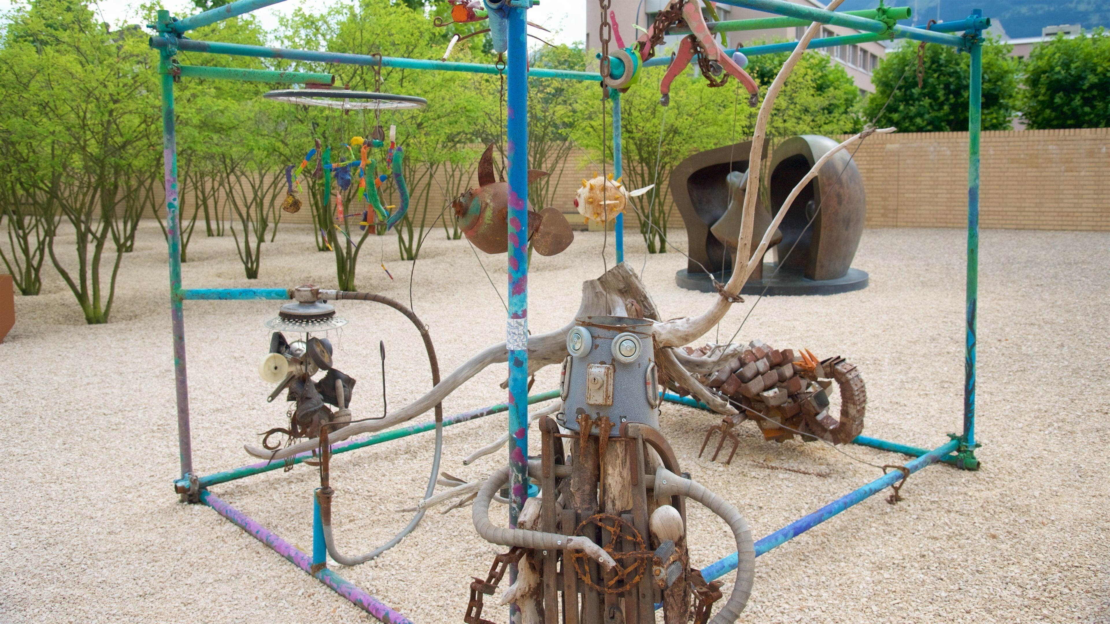 A sculpture of a mechanical creature with pipes and gears, set against a backdrop of trees and a brick wall.