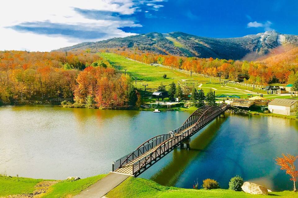 Un pont en bois enjambant un lac, avec des arbres aux couleurs automnales et une montagne en arrière-plan.