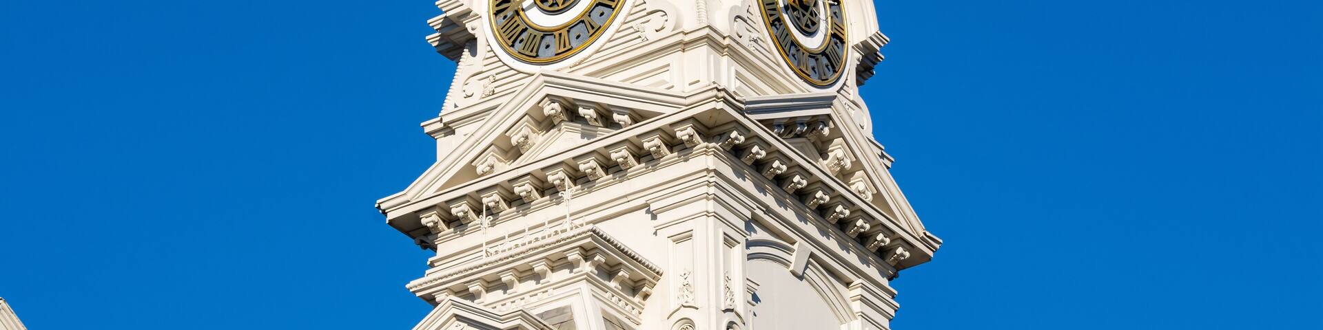 A white tower with two large clocks and intricate architectural details against a clear blue sky.
