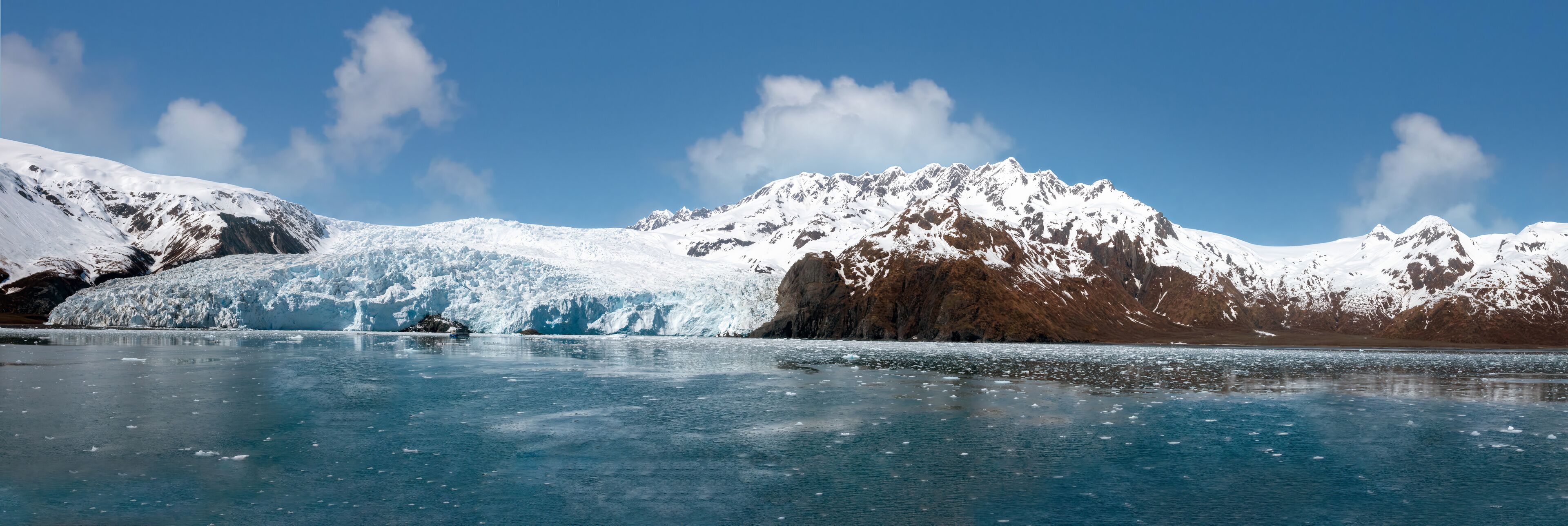 A glacier with a clear blue lake in front, surrounded by snow-covered mountains under a clear sky.