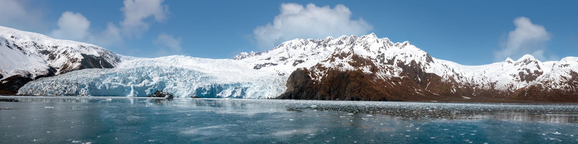 A glacier with a clear blue lake in front, surrounded by snow-covered mountains under a clear sky.