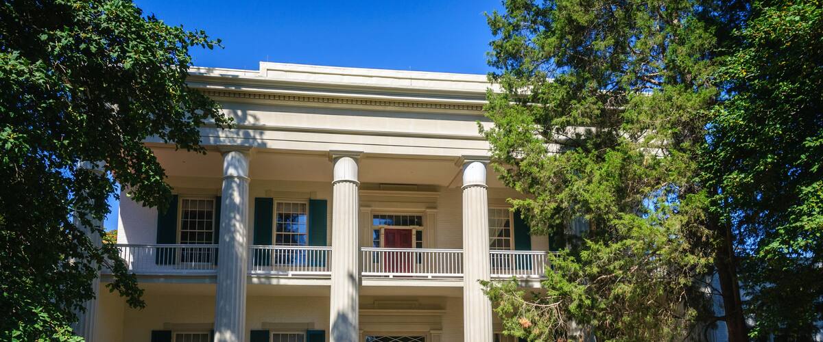 A historic building with white columns and green shutters, a balcony, and people sitting on benches in the foreground.