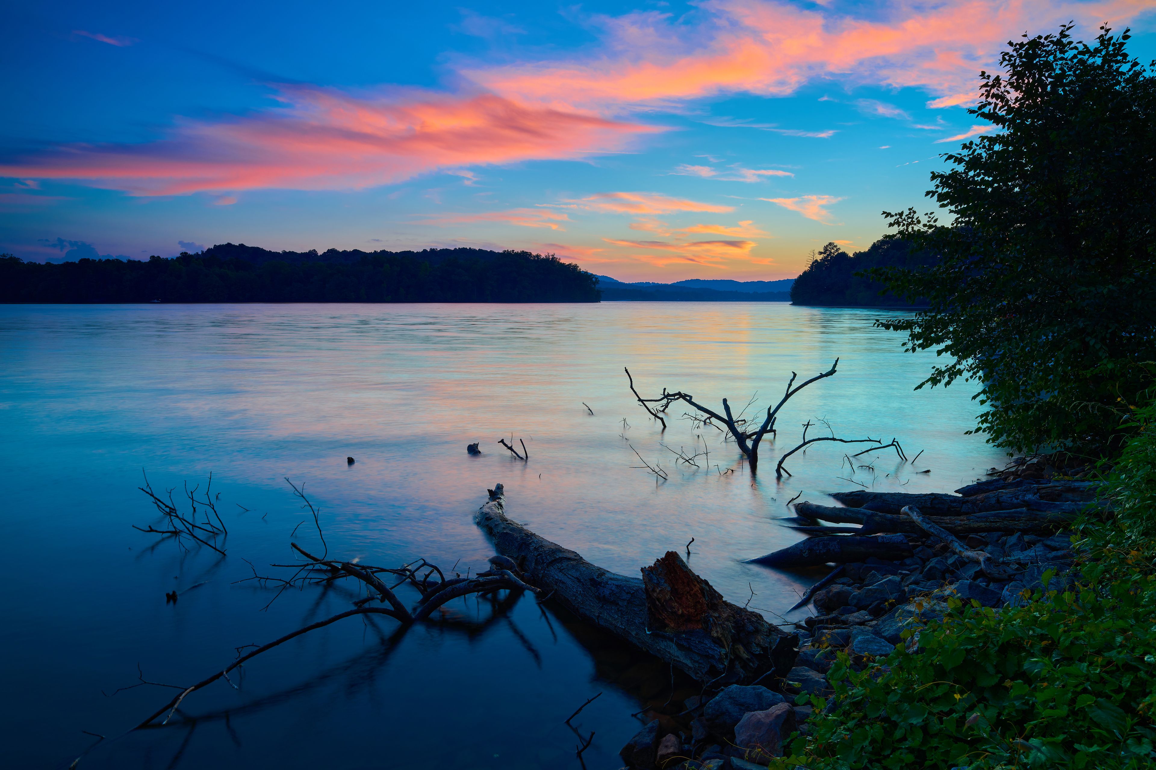 A calm lake at sunset with a colorful sky, surrounded by trees and rocks.