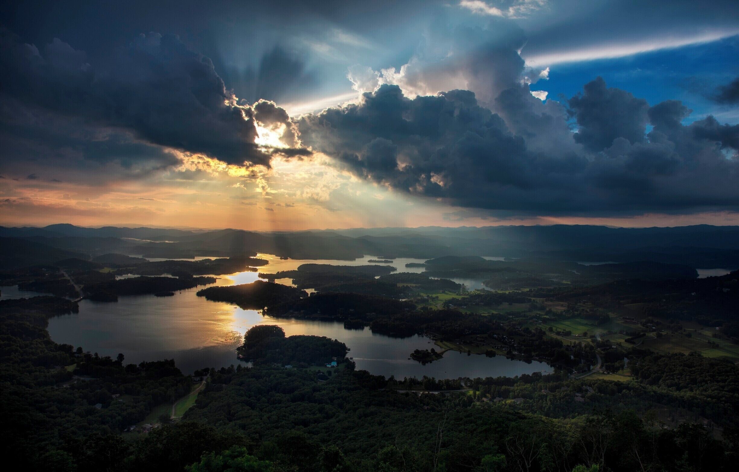 A landscape with a large body of water surrounded by forested hills and a dramatic sky with sunlight breaking through clouds.