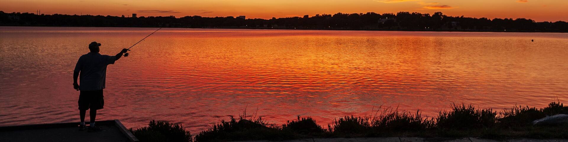 A person fishing at sunset by a calm lake with a vibrant sky.
