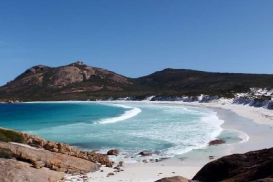 Un paisaje costero con una playa de arena, aguas turquesas cristalinas y una colina destacada al fondo.