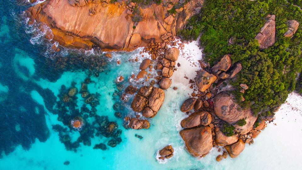 A coastal landscape with clear turquoise waters, large boulders, and lush greenery.