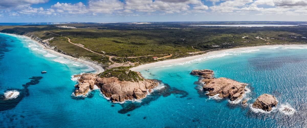 A coastal landscape with clear turquoise waters, rocky outcrops, and a winding road through a green forested area.
