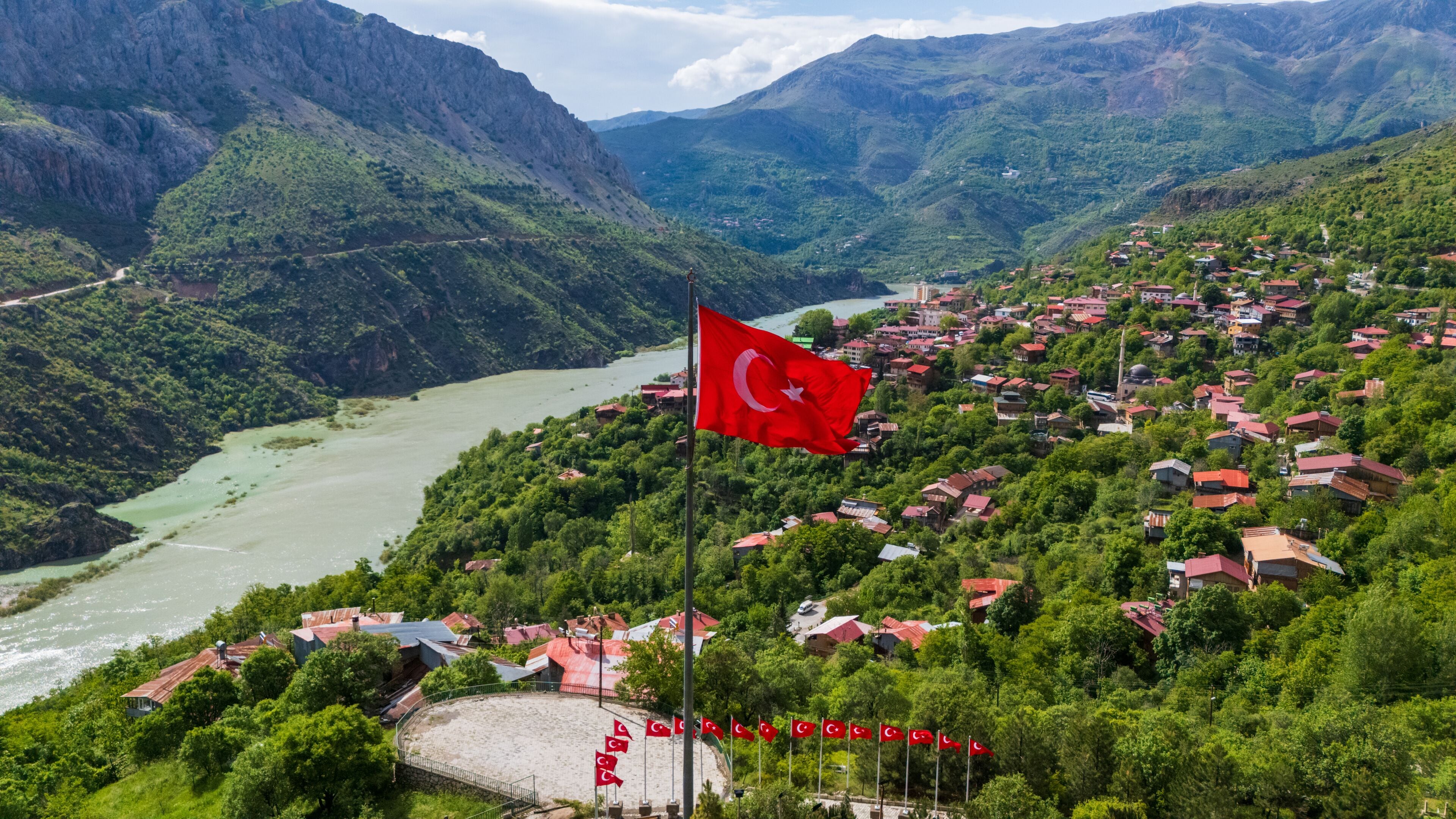 A mountainous landscape with a river, a village with red-roofed houses, and a Turkish flag.