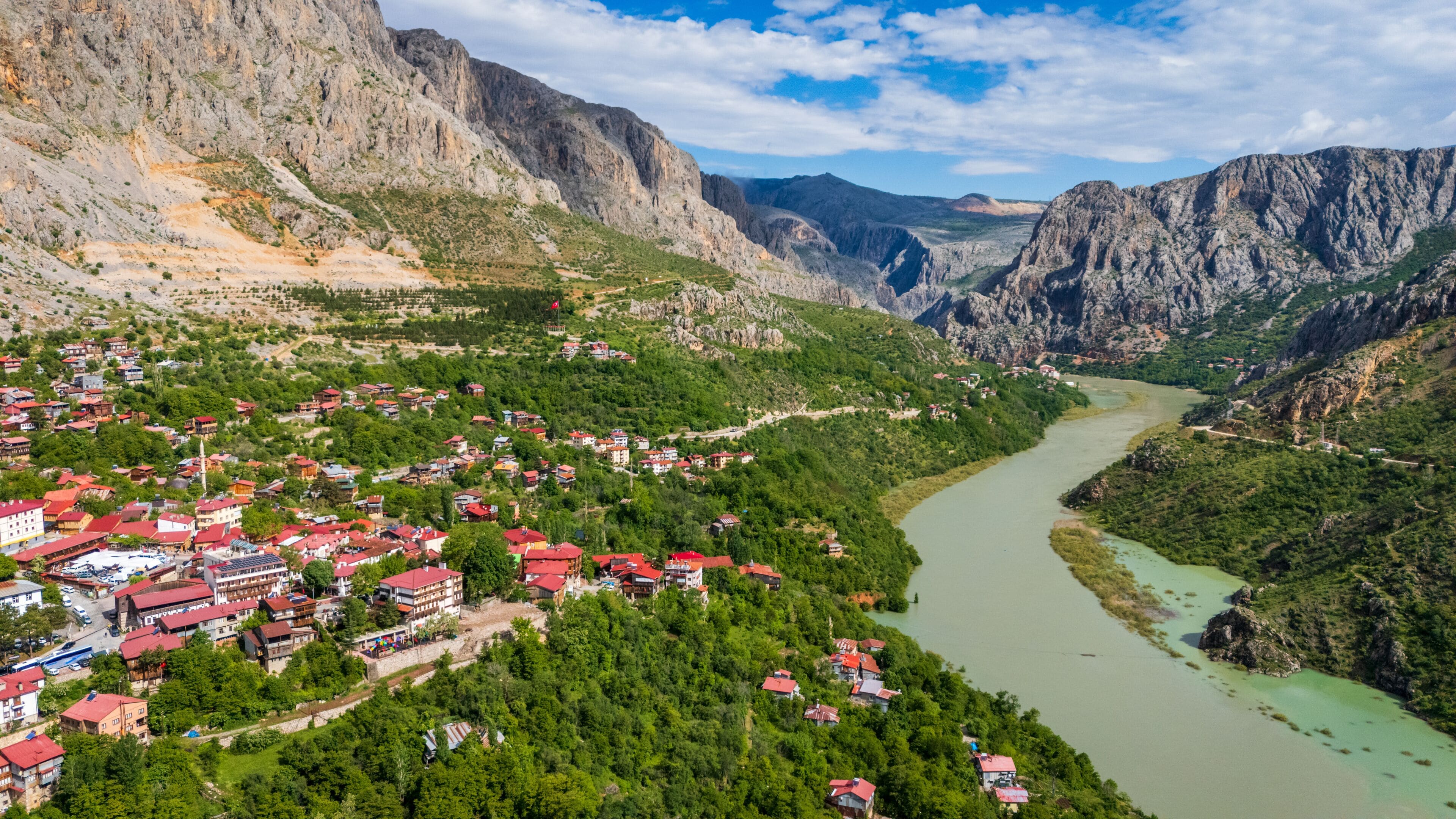A mountainous landscape with a river, a village with red-roofed houses, and a flag.