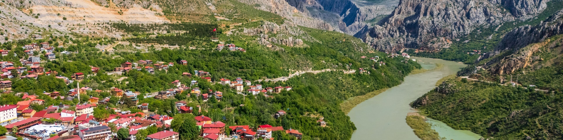 Un paisaje montañoso con un río, un pueblo con casas de techo rojo y una bandera.