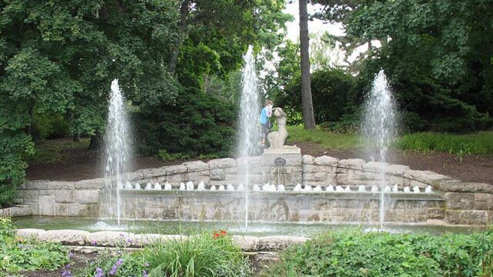 A stone fountain with three spouts surrounded by a garden with various plants and flowers.
