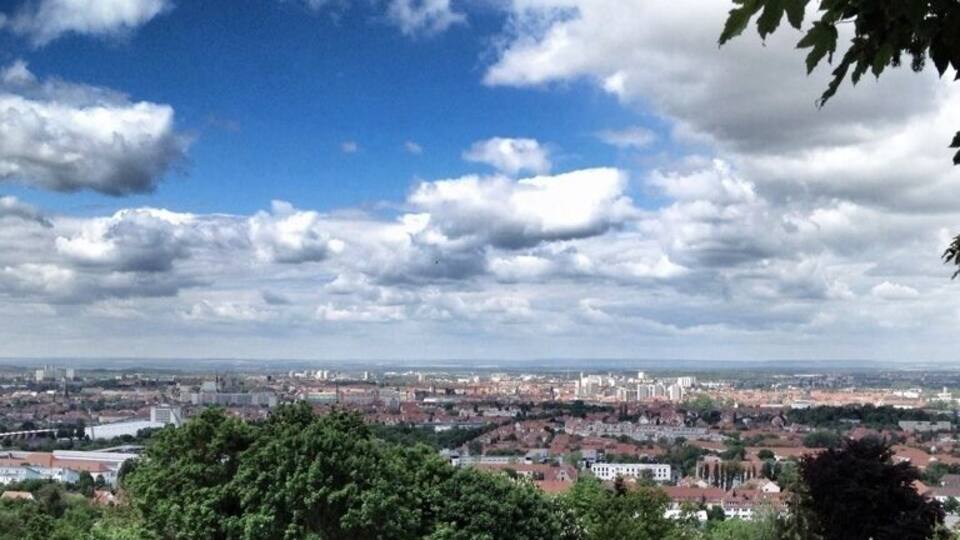 A cityscape with a hill in the foreground, a blue sky with clouds, and a mix of greenery and buildings.