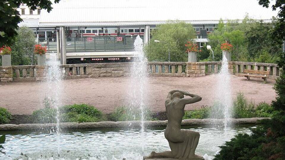 A fountain with water jets, a statue, and a train in the background.