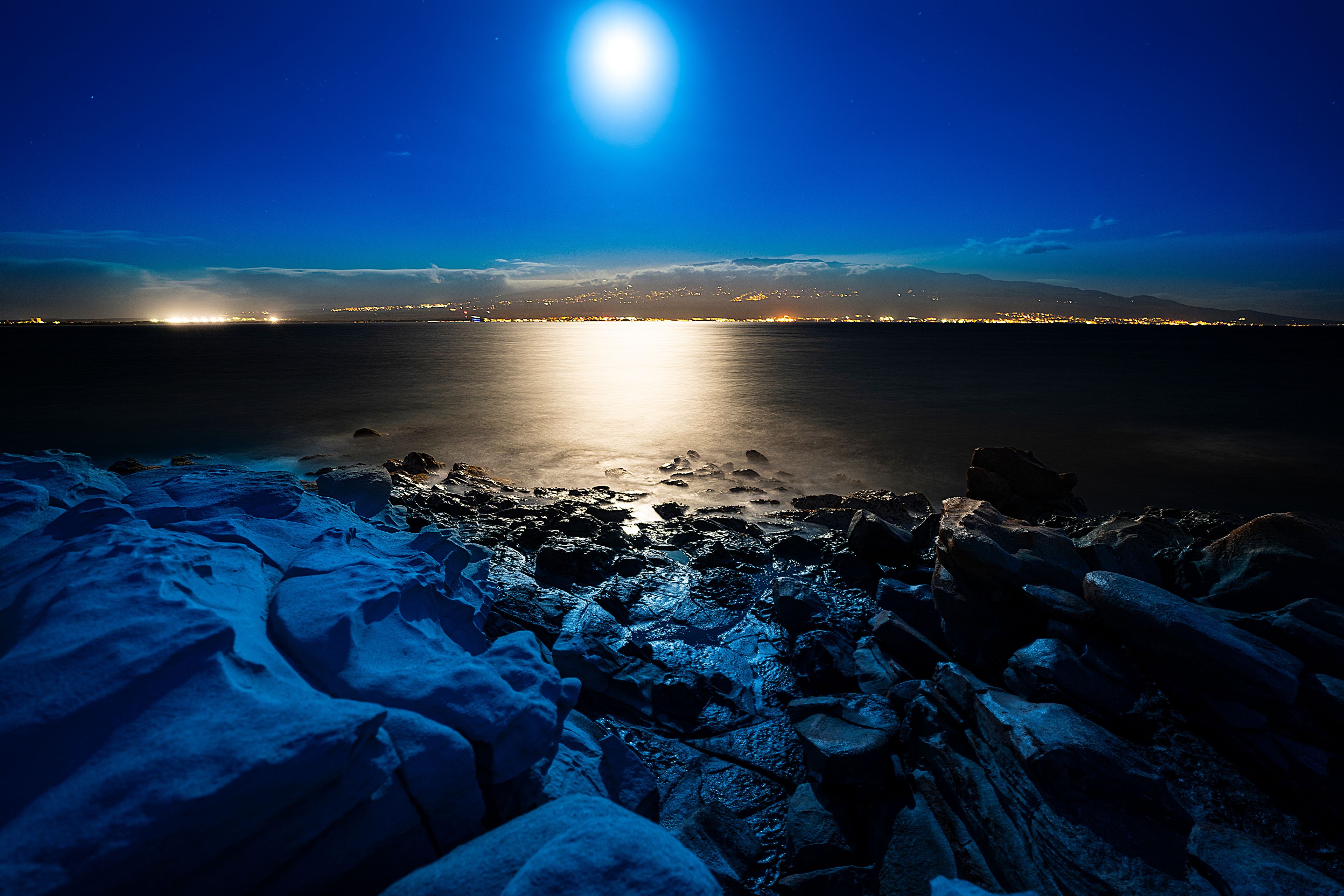 A nighttime coastal scene with a calm sea reflecting moonlight, rocky shoreline, and distant city lights.