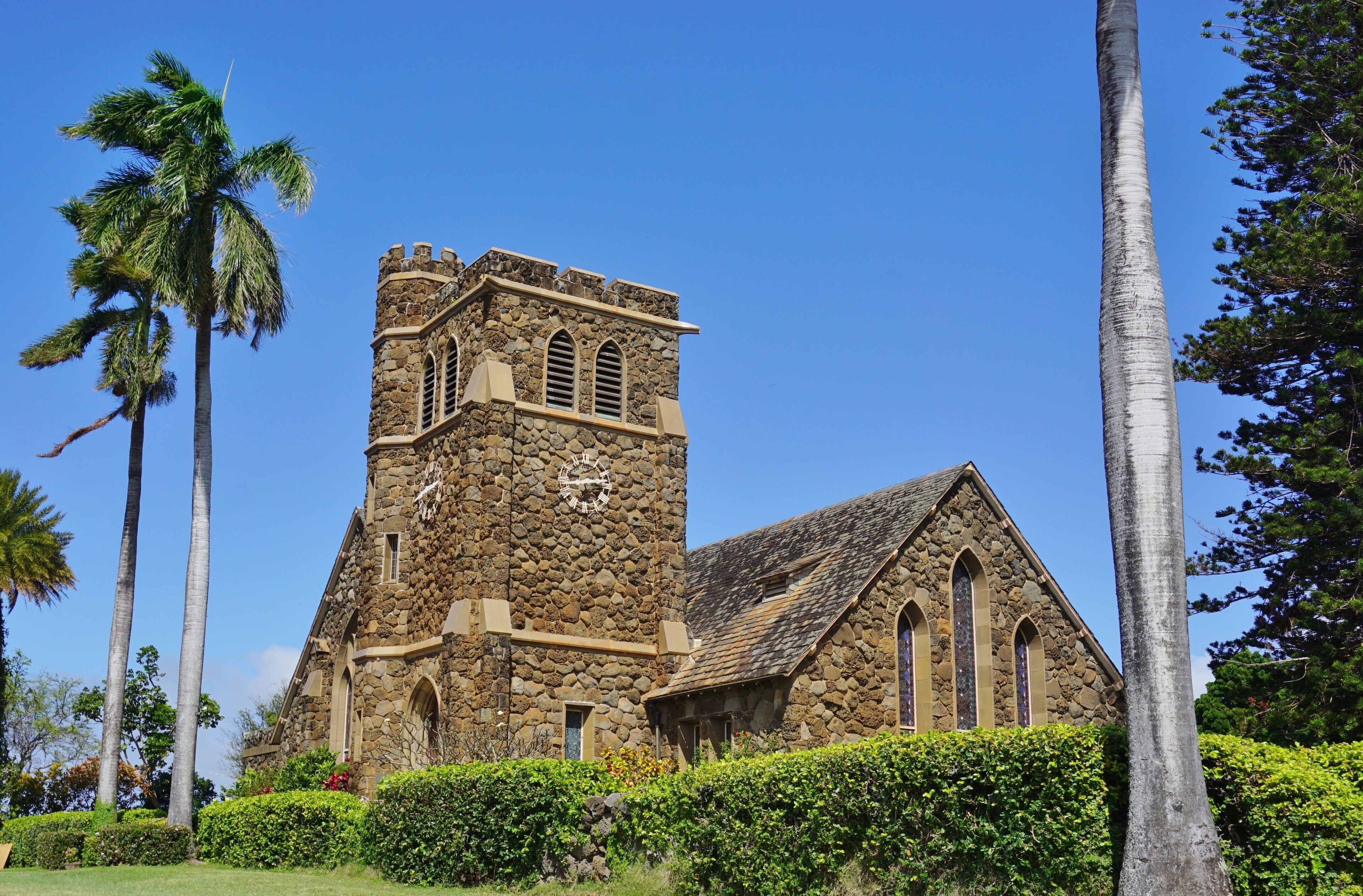 A stone church with a clock tower, surrounded by palm trees and greenery.
