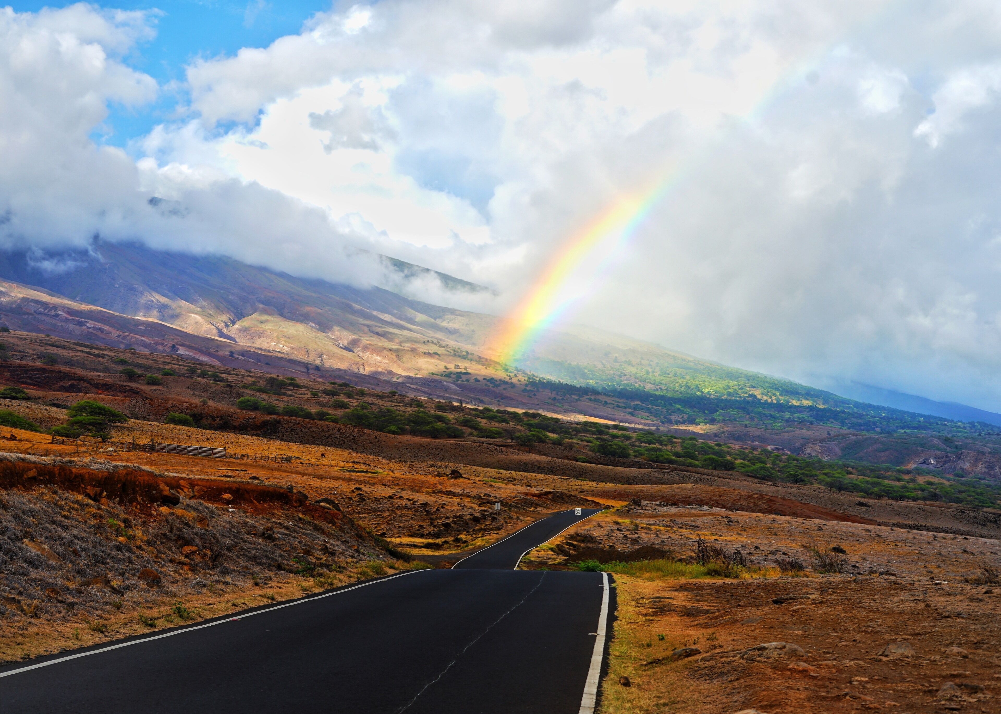 A winding road through a hilly landscape with a rainbow in the sky.