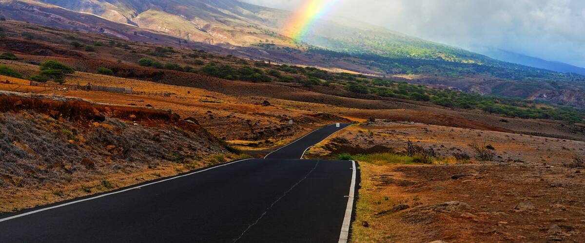 A winding road through a hilly landscape with a rainbow in the sky.