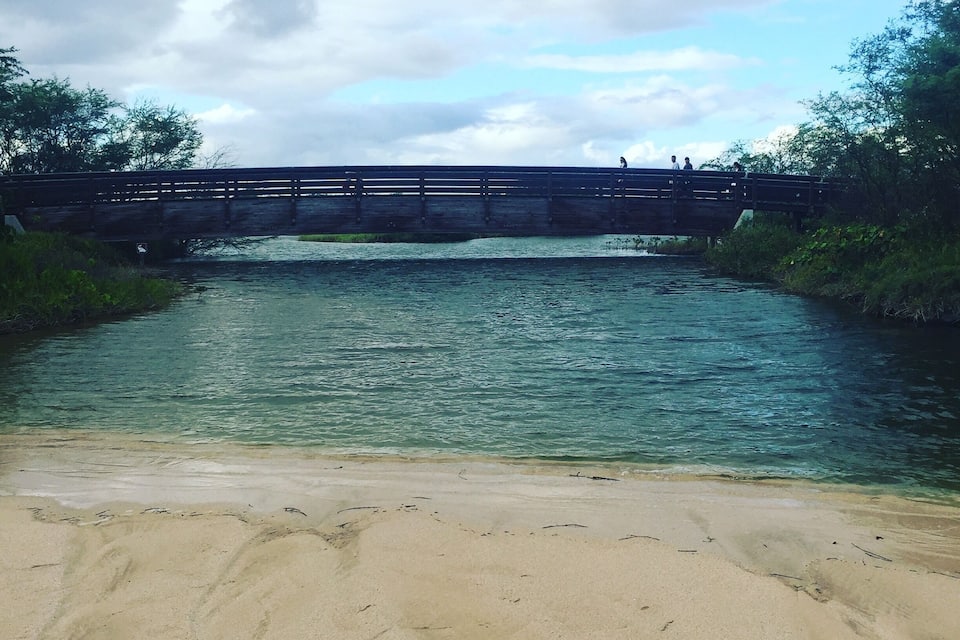 A sandy beach with a river, a wooden bridge, and a cloudy sky.