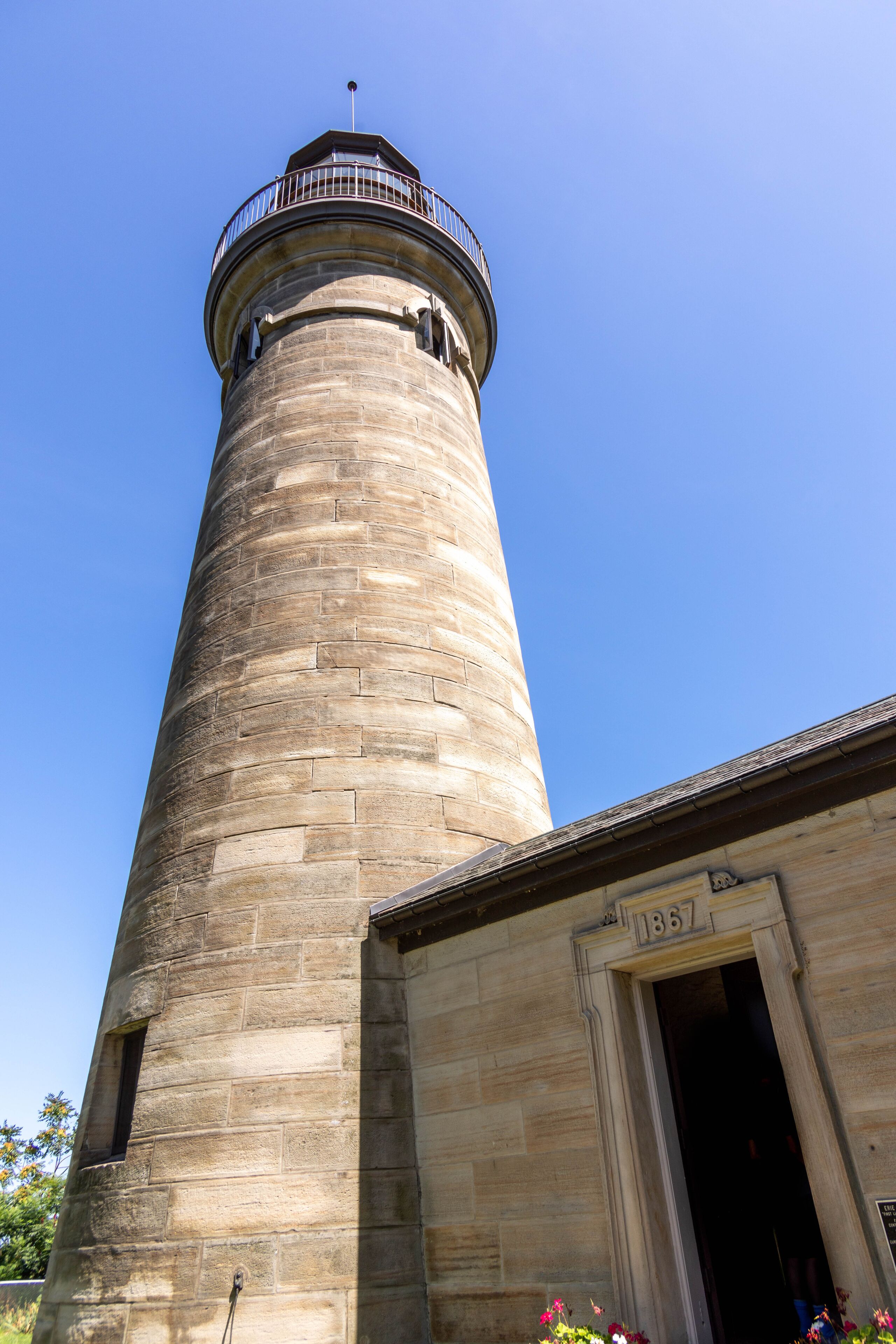 A tall stone lighthouse with a balcony and a plaque reading '1867' at its base.