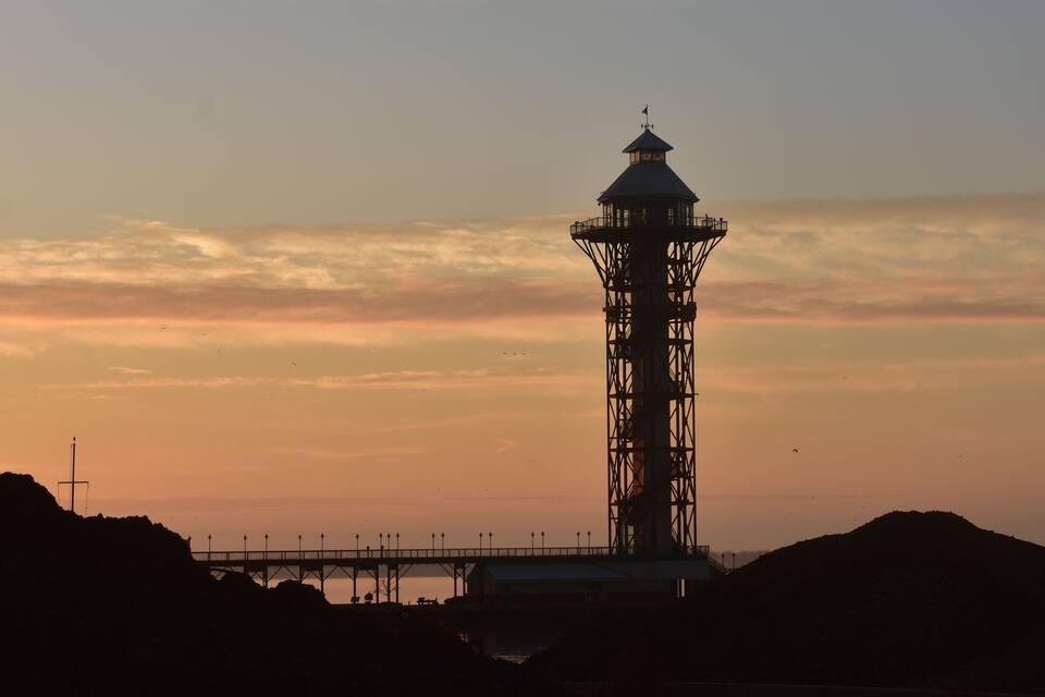 A lighthouse silhouette against a sunset sky, with a pier extending into the water.