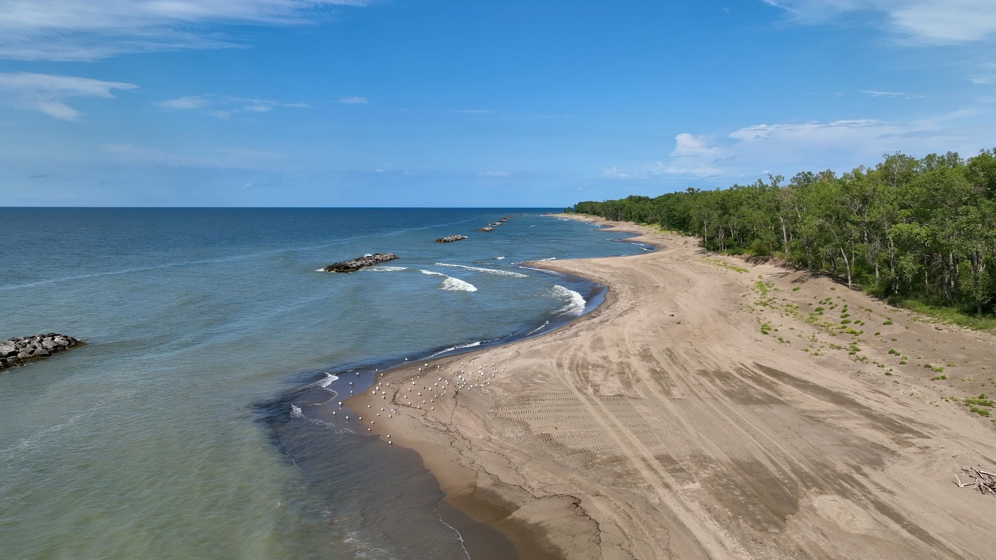 A sandy beach with clear blue waters, a rocky outcrop, and a line of trees.