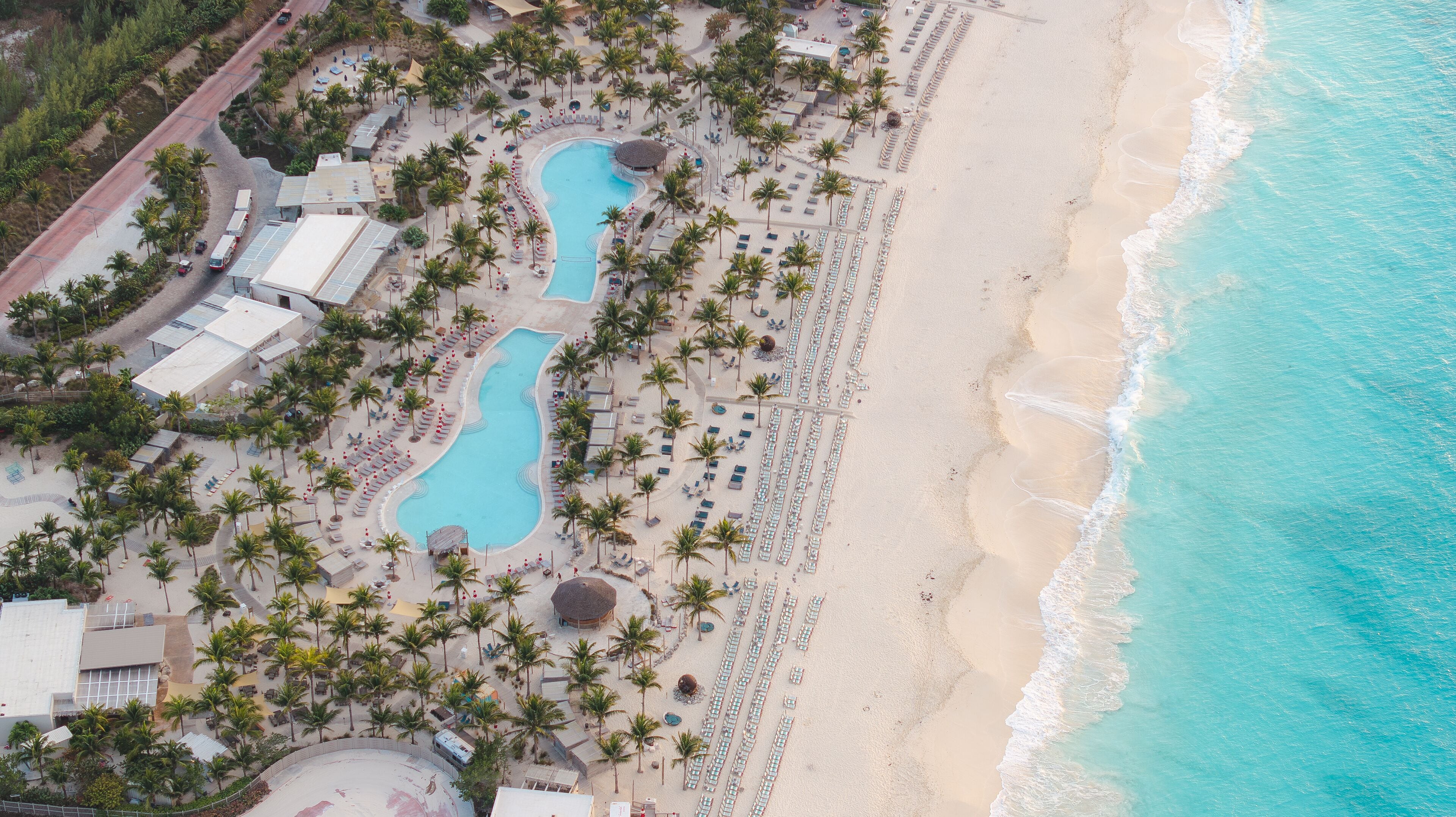 Aerial view of turquoise waters meet the sandy shore beside a resort with winding pools and palm trees, Alice Town, Bimini, The Bahamas.